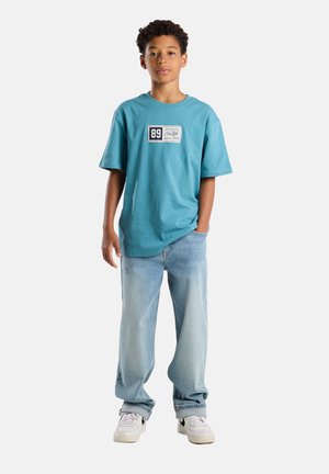 Türkises Baumwoll-T-Shirt mit einem rechteckigen Aufnäher mit der Beschriftung "89". Kombiniert mit ausgewaschenen blauen Jeans und weißen Sneakers mit schwarzen Akzenten.