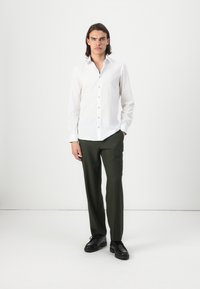White long-sleeve shirt with a button-up front, paired with dark green trousers and black shoes. Smooth fabric and fitted design.
