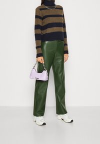 Persoon die een bruin en marineblauwe gestreepte sweater, groene leren broek, witte sneakers draagt en een kleine lavendelkleurige handtas met een zilveren kettinghengsel vasthoudt.