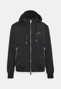 Musta bomber-takki hupulla, edessä vetoketju, sivuilla vetoketjulliset taskut ja resorit. Sisältää logotiedot rinnassa ja narut.