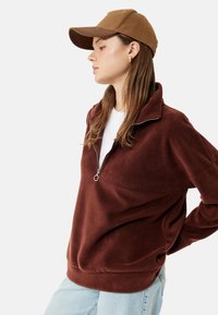 Brauner Fleece-Pullover mit einem halben Reißverschluss am Kragen, kombiniert mit einer hellbraunen Kappe. Der Pullover hat einen lockeren Schnitt und eine weiche Textur. Helle Jeans runden den Look ab.