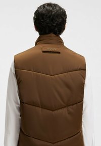 Braune Steppweste mit hohem Kragen und Zickzackstich-Muster. Verfügt über ein Markenlabel am Hals und glattes, glänzendes Material.