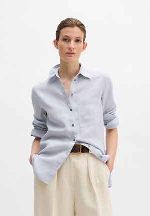 Kobieta ubrana w jasnoniebieską koszulę na guziki z podwiniętymi rękawami, beżowe spodnie z wysokim stanem i brązowy pasek, ręce w kieszeniach, neutralne tło.