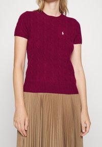 Fuchsiarosa kabelstickad kortärmad tröja med ribbad fåll och en liten logotyp på bröstet, kombinerad med en plisserad beige kjol.