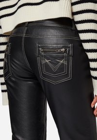 Zwarte leren broek met contrastkleurige witte stiksel. Heeft ritszakken op de achterkant en een strakke, aansluitende pasvorm. Gestreepte trui zichtbaar.