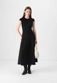 Robe maxi sans manches noire avec un corsage ajusté et une jupe évasée. Tissu lisse, design minimaliste, associée à des bottes noires à talons et un sac blanc.