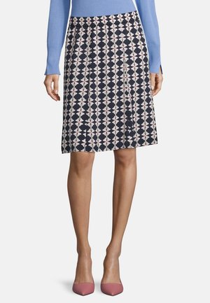 Femme portant une jupe noire longueur genoux avec un motif floral blanc et rose, un haut bleu à manches longues et des talons roses à bout pointu.