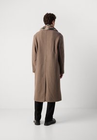 Abrigo de lana marrón con cuello de piel sintética, diseño recto, apertura en la parte trasera y dos bolsillos laterales, combinado con pantalones negros y zapatos oscuros.