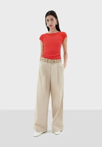 Camiseta roja ajustada combinada con pantalones anchos beige y un cinturón de estampado. El modelo lleva zapatos blancos y tiene el cabello negro largo y liso.