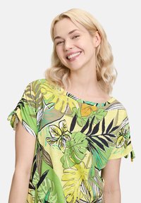 Zielony top w kwiaty wykonany z lekkiego materiału. Posiada nadruk tropikalnych liści, krótkie rękawy z wiązaniami i luźny fason.