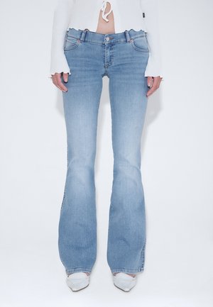 Kvinde iført lyseblå jeans med høj talje og vidde ben, hvide spidse sko og en kort hvid top med lange ærmer og bindedetalje foran.