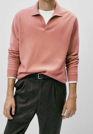 Mężczyzna w łososiowo-różowym, długim swetrze dzianinowym na białej koszuli, w połączeniu z ciemnoszarymi plisowanymi spodniami i czarnym paskiem.