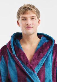 Pluszowy szlafrok w naprzemiennych fioletowych i turkusowych paskach, z kołnierzem szalowym i miękką teksturą. Idealny do komfortu i relaksu.