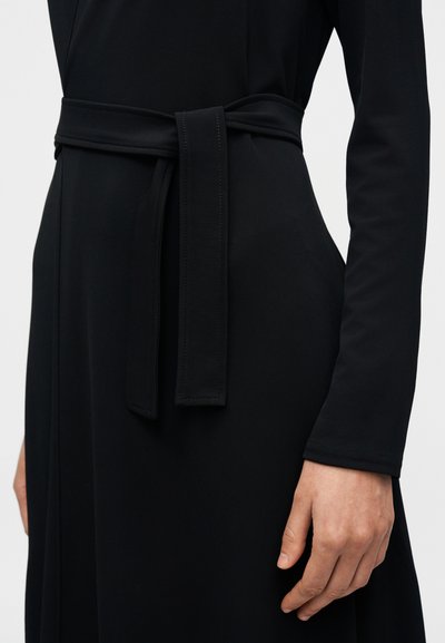 Μαύρο φόρεμα με μακριά μανίκια, διαθέτει δέσιμο στη ζώνη στη μέση. Ομαλό, υφασμάτινο ύφασμα με διακριτικές ραφές στη ζώνη.