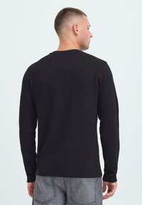 Črna, dolgimi rokavi srajca iz teksturirane tkanine, ima okroglo ovratnik in ožji kroj. Prikazana je zadaj, v kombinaciji s sivimi hlačami.