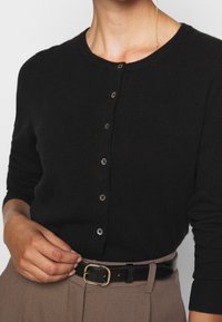 Cardigan nero con scollo rotondo, cinque bottoni sul davanti e maniche lunghe. Abbinato a una cintura marrone con fibbia dorata.