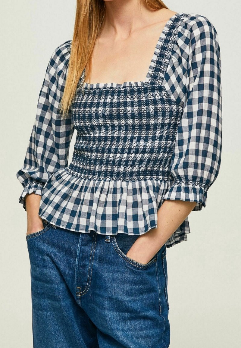 Blusa a quadri blu e bianchi con corpetto arricciato, scollatura quadrata e orlo a volant. Maniche lunghe a sbuffo con polsini elastici.