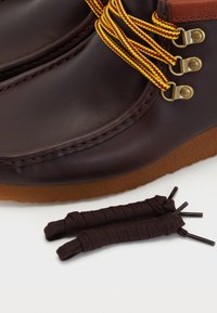 Botas de cuero marrón con cordones tejidos en amarillo y rojo, ojales dorados y una suela de goma color beige; incluye cordones marrones de bota al lado.