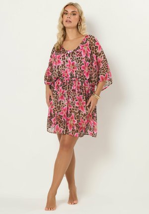 Femme debout pieds nus portant une robe courte et ample à imprimé floral rose et léopard, mains posées sur les hanches, fond neutre.