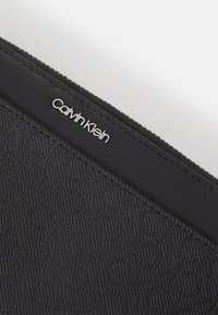 Black zippered pouch featuring a textured pattern and a silver "Calvin Klein" logo on the top edge. Smooth material with a matte finish.