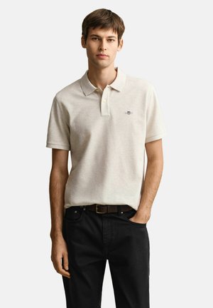 Junger Mann mit beige-farbenem Kurzarm-Poloshirt und schwarzen Hosen mit braunem Gürtel, steht mit einer Hand in der Tasche.