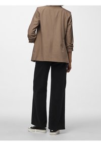 Hnědý tailored blazer s třičtvrtečními rukávy, kombinovaný s černými širokými kalhotami a černými teniskami s bílou podrážkou. Hladká textura, minimální design.