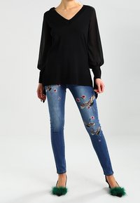 Schwarzer V-Ausschnitt-Pullover mit durchsichtigen Ballonärmeln, kombiniert mit blauen Jeans, die mit Blumen- und Vogel-Stickereien verziert sind, sowie detaillierten grünen Pelz-Absätzen.