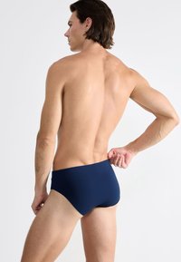 Marineblå svømmebukser vises fra ryggen. De har en glat tekstur og et enkelt, figursyet design med en beskeden talje.