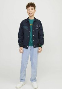 Giubbotto bomber navy con bottoni bianchi, dettagli verdi e toppe ricamate. Abbinato a una maglietta verde e jeans azzurri chiari. Sneakers bianche.