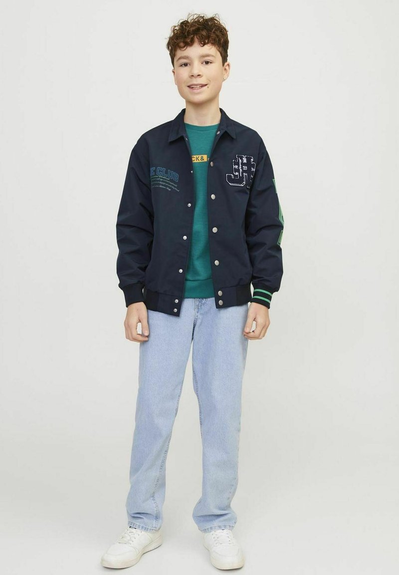 Giubbotto bomber navy con bottoni bianchi, dettagli verdi e toppe ricamate. Abbinato a una maglietta verde e jeans azzurri chiari. Sneakers bianche.
