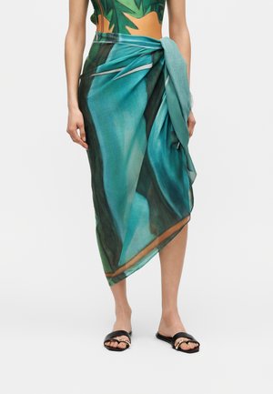 Femeie care poartă un sarong cu imprimeu tropical verde și albastru legat în talie, asortat cu sandale negre plate, cu degetele goale și accente aurii.