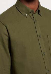 Camicia button-up verde oliva scuro con colletto abbottonato e taschino visibile su una persona con tonalità di pelle scura.