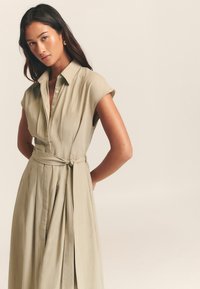 Haljina košulja boje kaki s kratkim, podvrnutim rukavima, gumbima sprijeda, povezom u struku i detaljem plisirane suknje. Glatka tekstura, minimalistički dizajn.