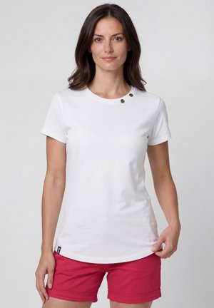 Frau mit dunklen Haaren, die ein weißes kurzärmeliges Hemd mit zwei Knöpfen am Kragen und rote hochgekrempelte Shorts trägt.