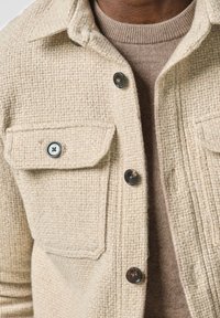 Giacca camicia beige tessurizzata con un motivo a quadri, dotata di due tasche frontali e bottoni scuri, indossata sopra un maglione marrone chiaro.