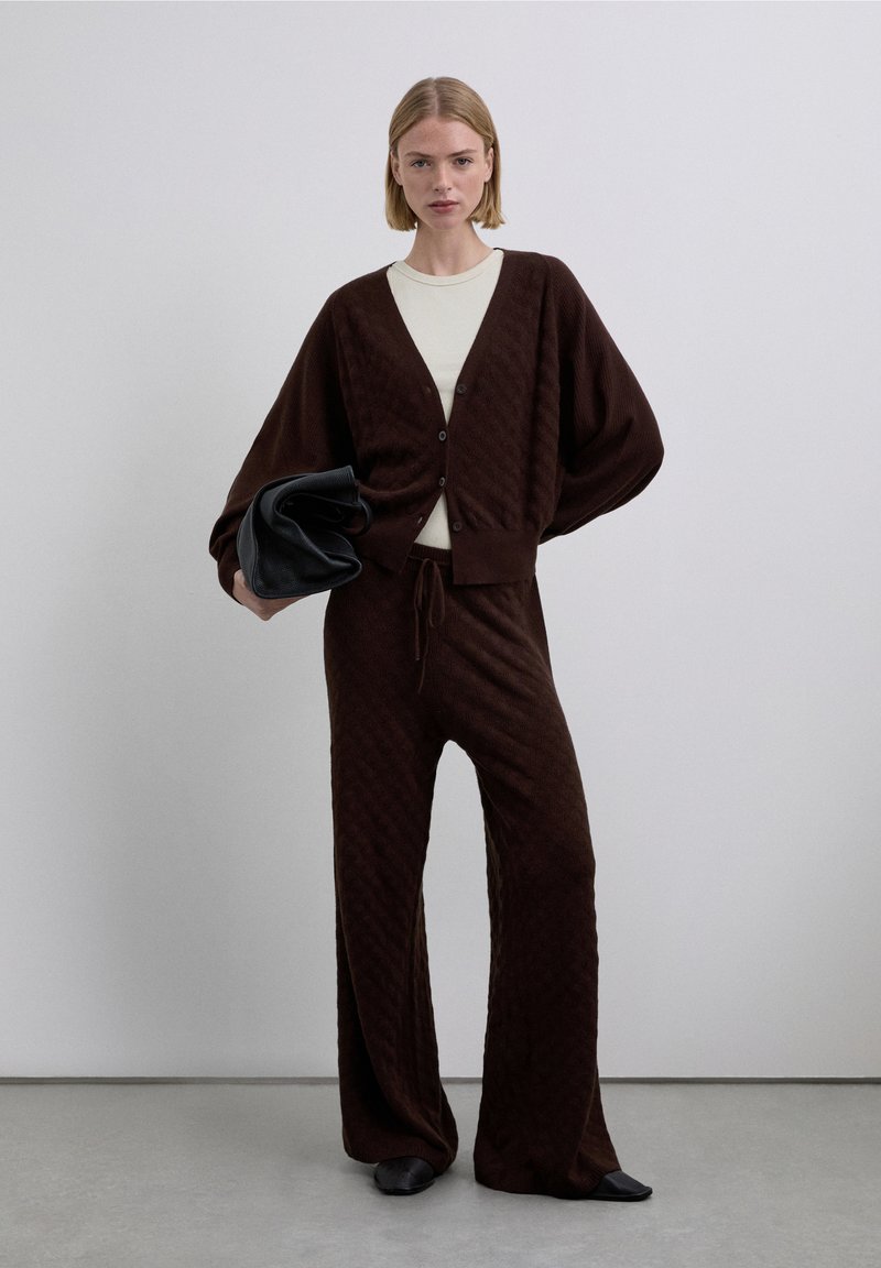 Cárdigan marrón tejido con un patrón de diamantes, combinado con pantalones sueltos a juego. El conjunto incluye una camiseta crema y un bolso de mano negro.