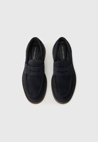 Une paire de mocassins en daim noir avec des détails de couture et des semelles intérieures marquées, vue de dessus sur un fond neutre.