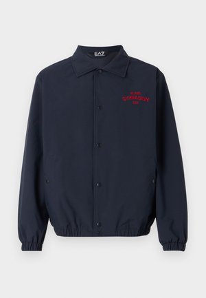 Giacca a vento blu navy realizzata in tessuto morbido, con chiusura a bottoni, polsini elasticizzati e ricamo rosso "GYMNASIUM EAZ" sul petto.