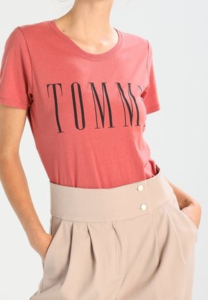 Vrouw draagt een rood T-shirt met korte mouwen met "TOMMY" in grote zwarte letters, ingestopt in een beige broek met hoge taille en knopen.