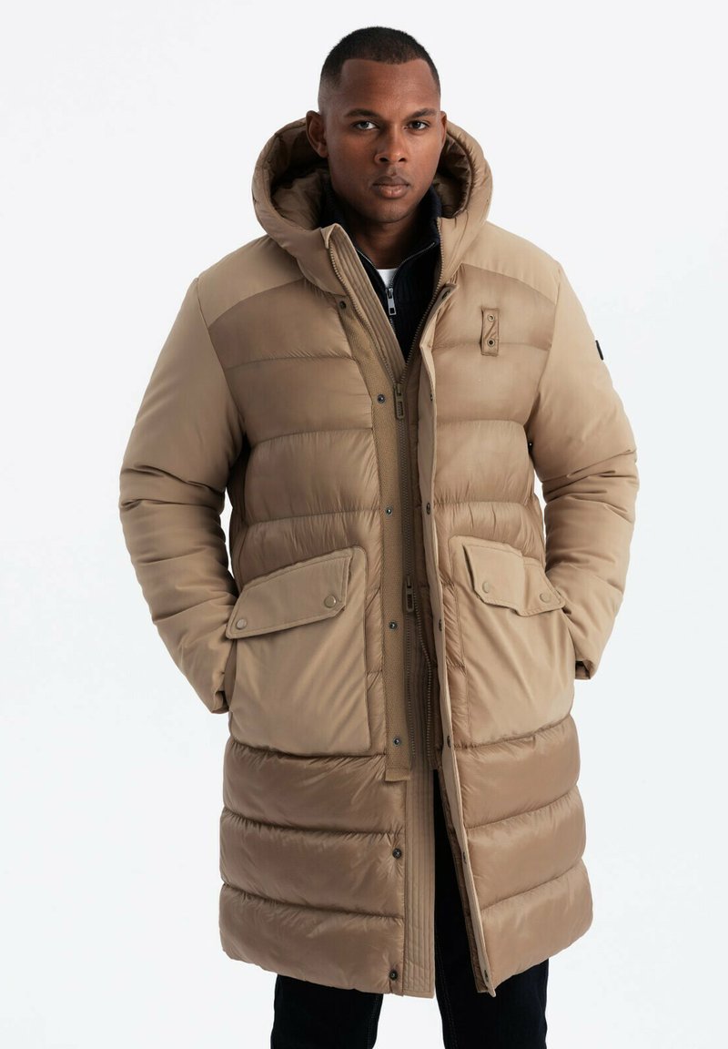 Beige gepolsterter Wintermantel mit Kapuze, ausgestattet mit Reißverschluss und Druckknopfverschluss, zwei frontalen Taschen und einem gesteppten Design.