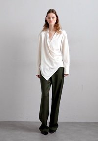 Blusa creme com mangas compridas e decote em V, combinada com calças verde-escuras justas e sapatos pretos de bico. Textura de tecido suave.