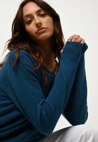Junge Frau mit langen braunen Haaren, die einen blauen Pullover und weiße Hose trägt, die Hände vor ihrem Gesicht gefaltet und in die Kamera schauend.