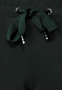 Mörkgröna joggingbyxor med en texturerad tyg, med dragsko i midjan och metallbeslag samt öljetter. Närbild på midjan.