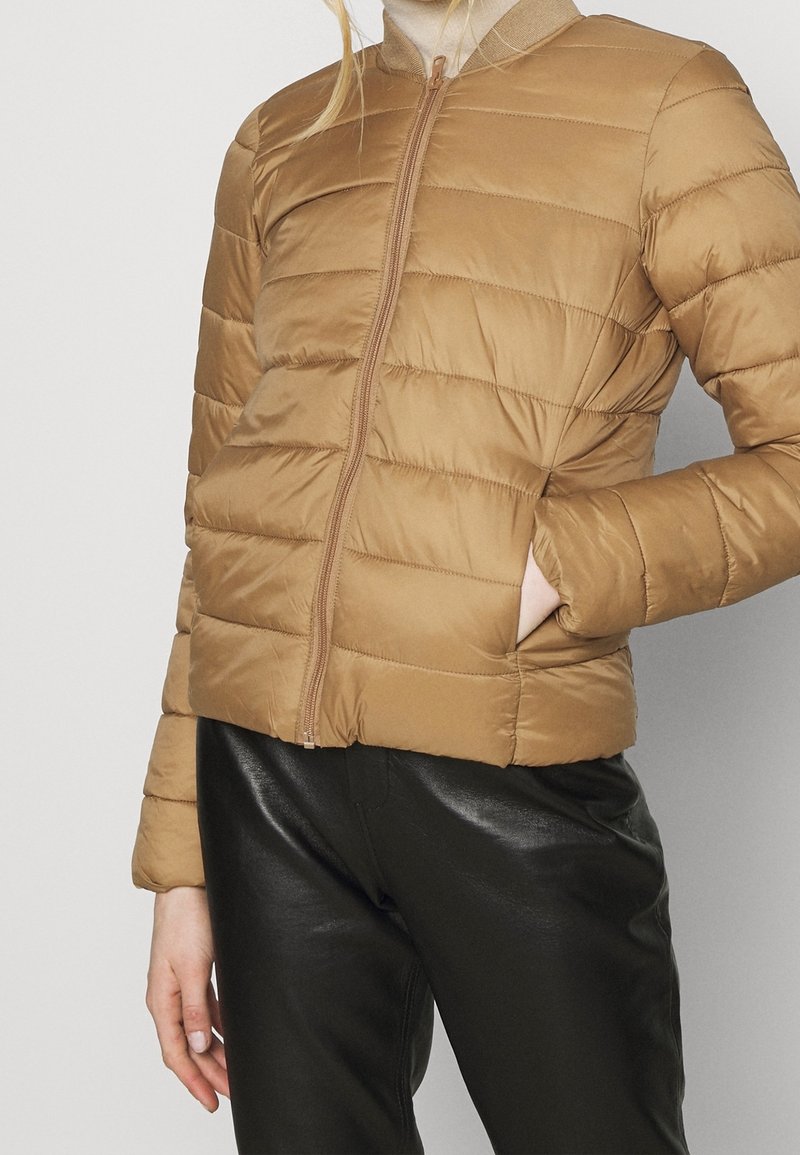 Kvinna som har på sig en tan-färgad vadderad zip-up jacka med sidofickor och svarta läderbyxor, handen i jackfickan.