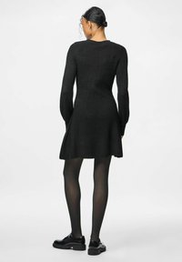 Abito nero lavorato a maglia con maniche lunghe, corpino aderente, gonna svasata e texture a coste. Abbinato a collant neri opachi e scarpe.