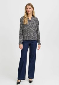 Schwarz-weiß gemusterte Bluse mit langen Ärmeln, V-Ausschnitt, gerafften Manschetten, kombiniert mit dunkelblauen Schlagjeans und schwarzen spitzen Schuhen.