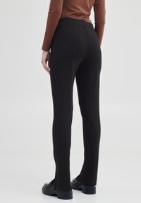 Leggings noirs avec une silhouette ajustée et une texture lisse, dotés d'une taille haute et de détails de couture sur le côté, associés à des chaussures noires.