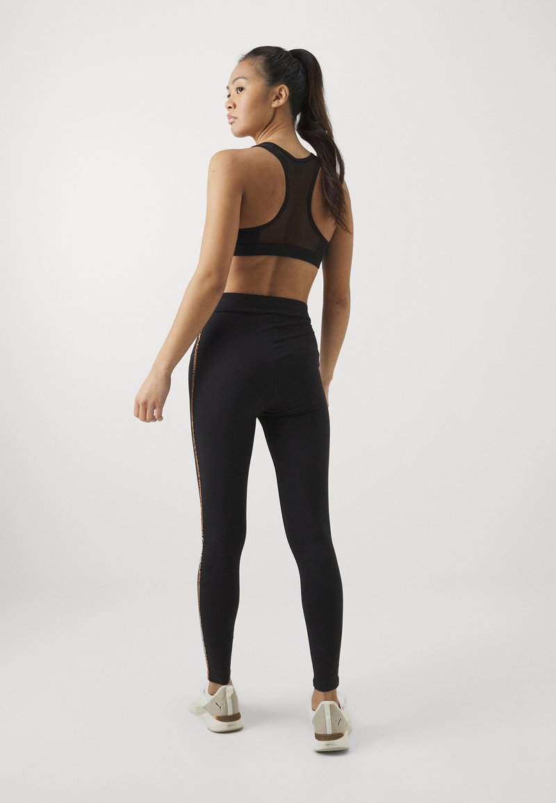Sujetador deportivo negro con detalles de malla, combinado con leggins negros de cintura alta que tienen un acento lateral. El modelo lleva zapatillas deportivas blancas.