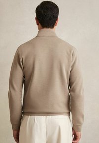 Beige huppari, jossa on kaulus, pitkät hihat ja sileä rakenne. Vaate on yhdistetty vaaleisiin housuihin.