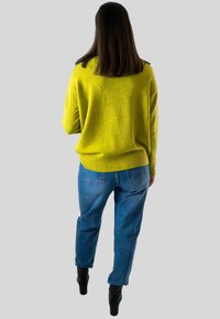 Vrouw met steil bruin haar, gekleed in een heldergele trui, blauwe jeans en zwarte hakken, staande met haar gezicht weg van een effen achtergrond.